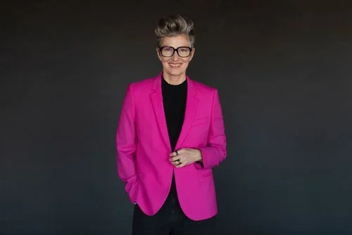 Jeanette Bronée wearing a pink blazer and black shirt with glasses, standing against a dark background.