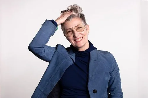 Person smiling and touching their hair, wearing glasses and a blue blazer.