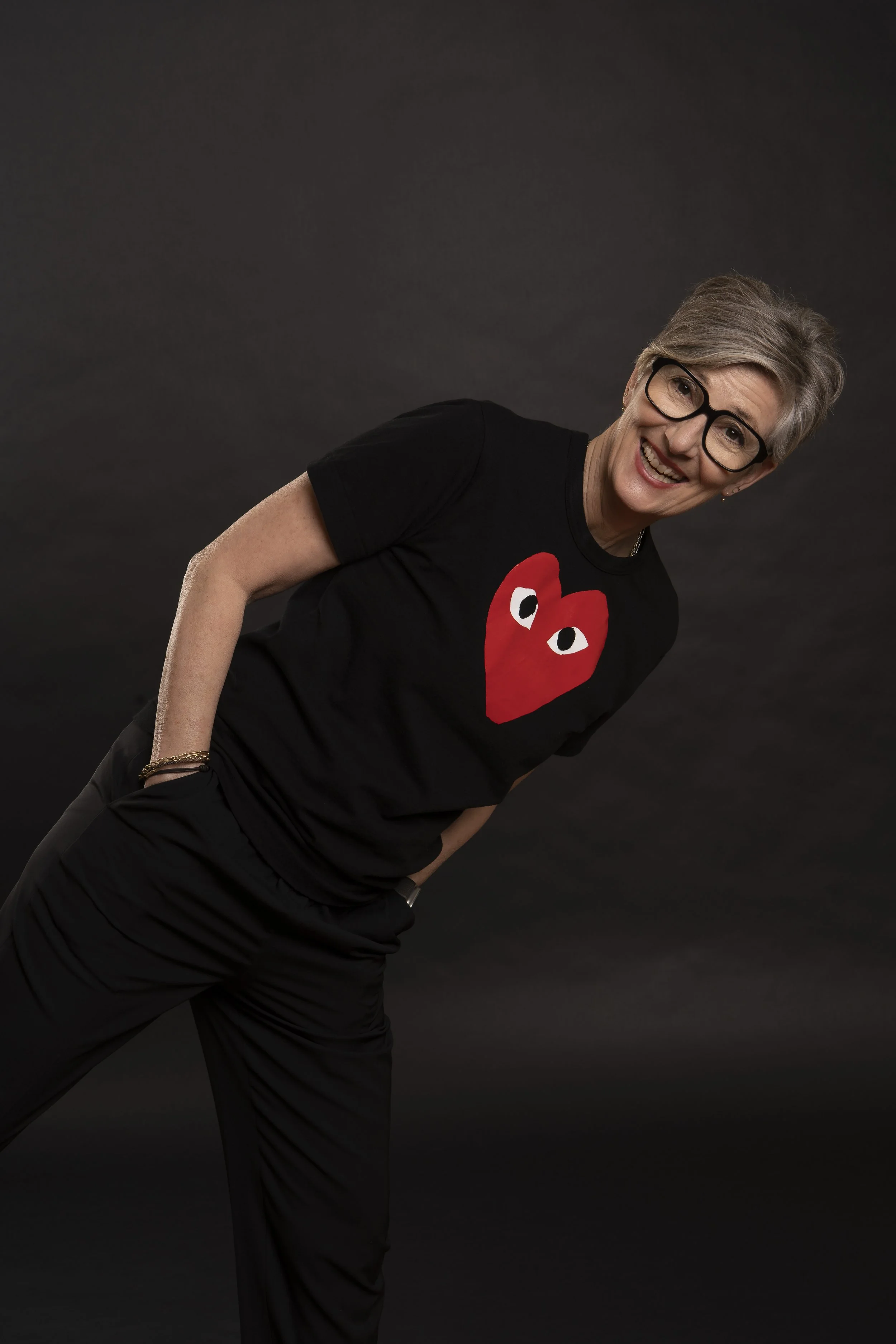 Jeanette Bronée, with short gray hair, glasses, and a black T-shirt featuring a red heart with eyes, smiling and posing against a dark background.