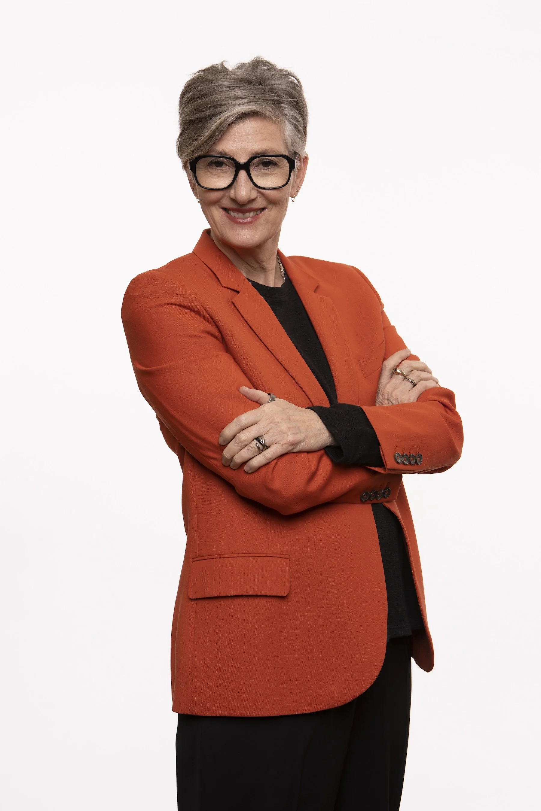 Jeanette Bronée with short gray hair, glasses, and earrings smiling with crossed arms, wearing an orange blazer over a black top against a white background.
