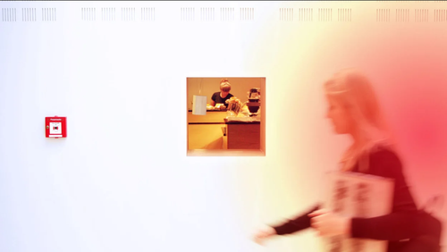 Blurry person passing by a translucent wall with a small window showing two people behind a counter.