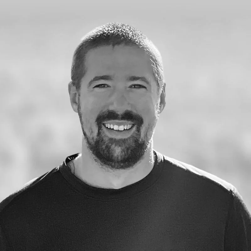 Black and white photo of a smiling man named John Keller with short hair and a beard, wearing a dark t-shirt, outdoors with a blurred background.