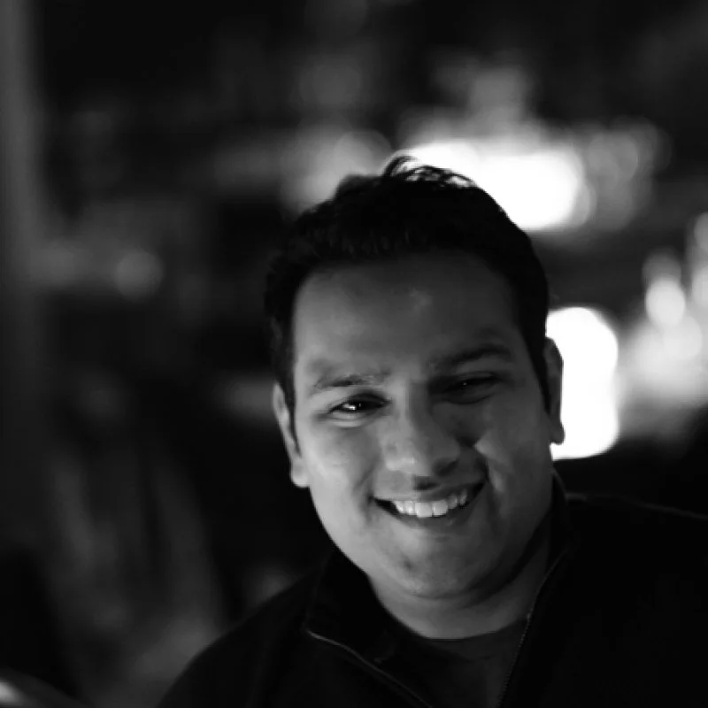 A smiling man named Saahil Jain, with dark hair in a black jacket, outdoors at night with blurred city lights in the background.
