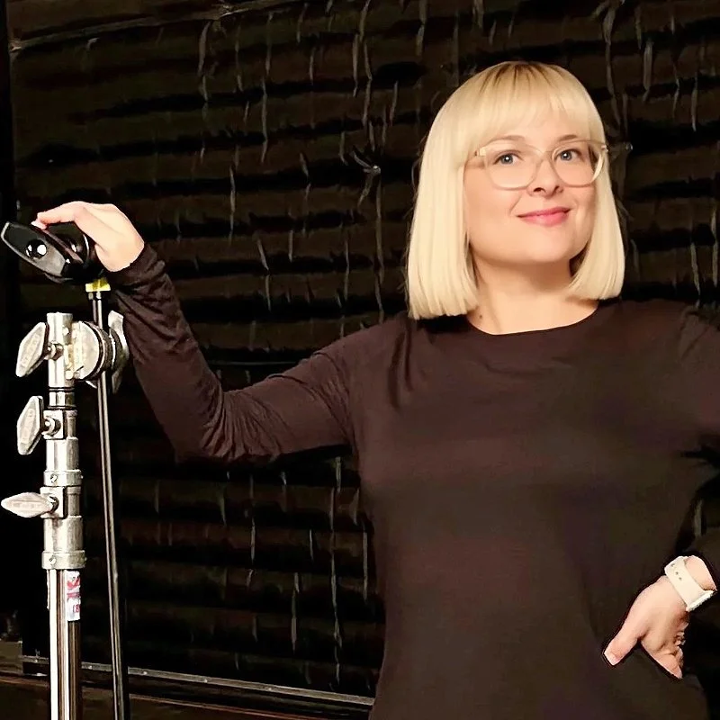 A woman named Jessica Miller, with blond hair, glasses, and a black long-sleeve shirt standing in front of a black background with vertical lines, holding a lighting stand with her right hand and smiling.