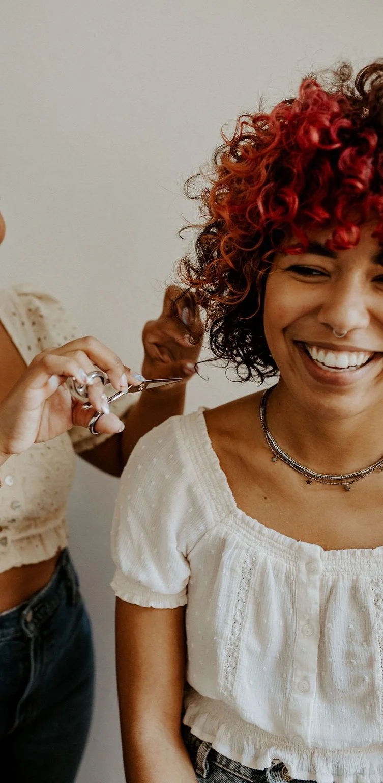Woman getting a haircut, smiling, with curly red hair and wearing a white blouse.