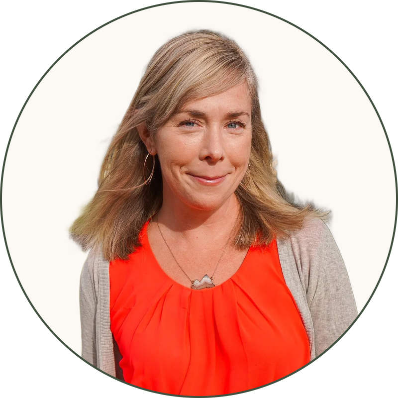 A woman with blonde hair wearing a red top and black pants standing outdoors next to a large tree, with a park in the background.