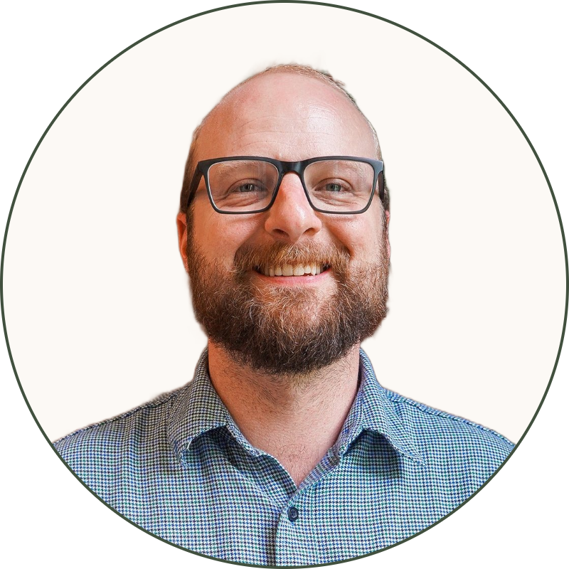A man with glasses, a beard, and a mustache is smiling while standing in front of a red brick wall and green plant.