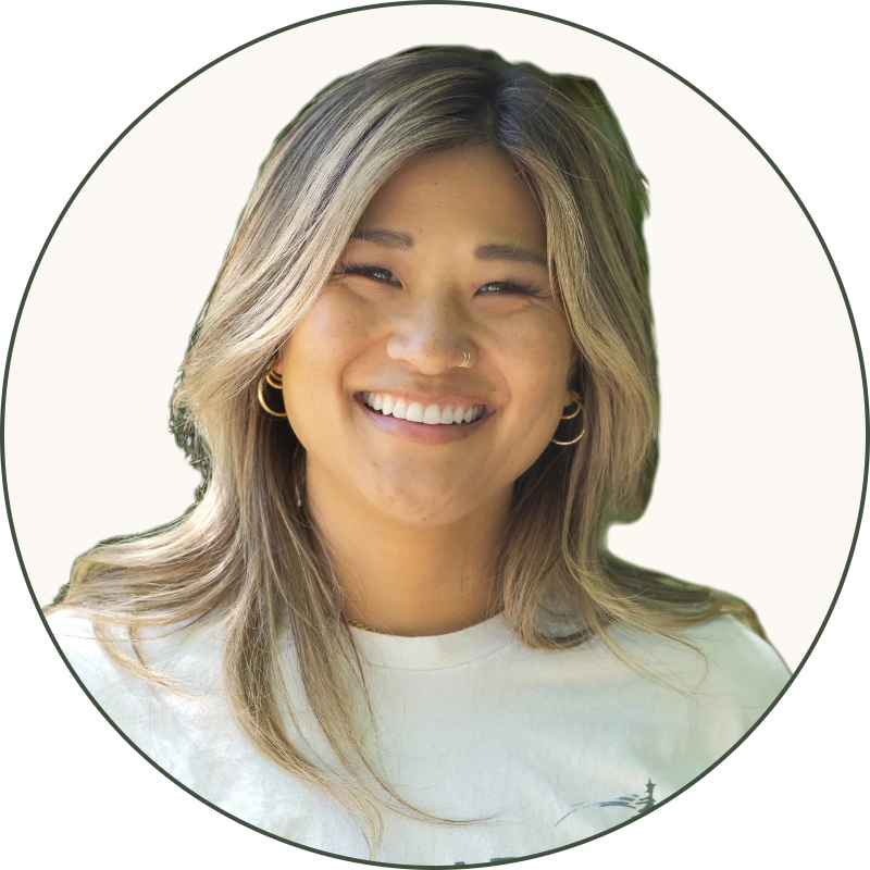 A smiling woman with blonde hair wearing earrings and a white T-shirt with the words 'Sustainable Northwest' and a pine tree logo, outdoors with green foliage in the background.