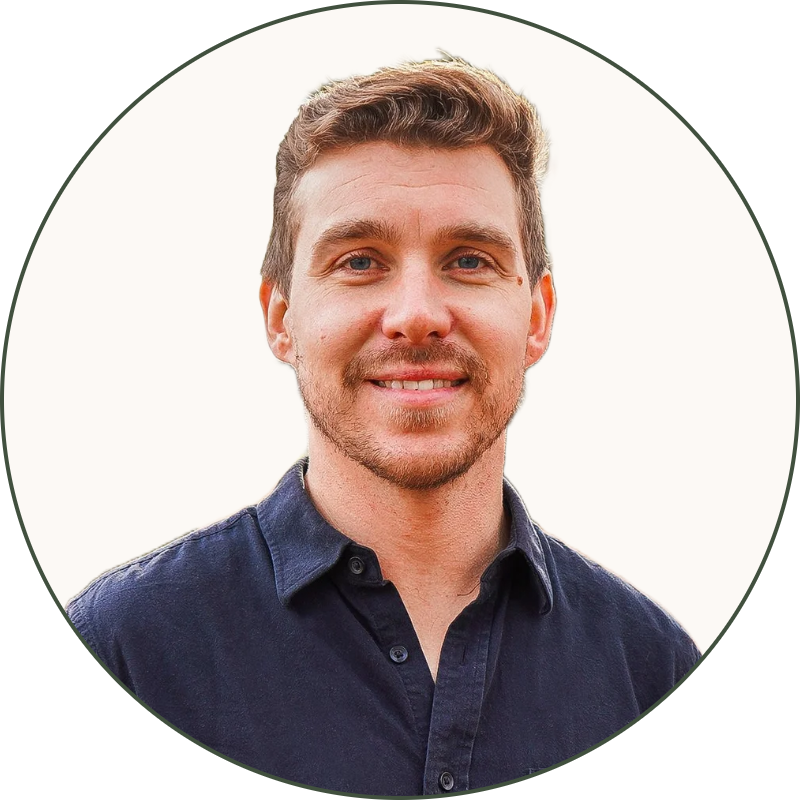 A man with brown hair and a beard standing outdoors in a park with fallen leaves, trees, and a street in the background during autumn. He is wearing a navy blue button-up shirt and khaki pants.