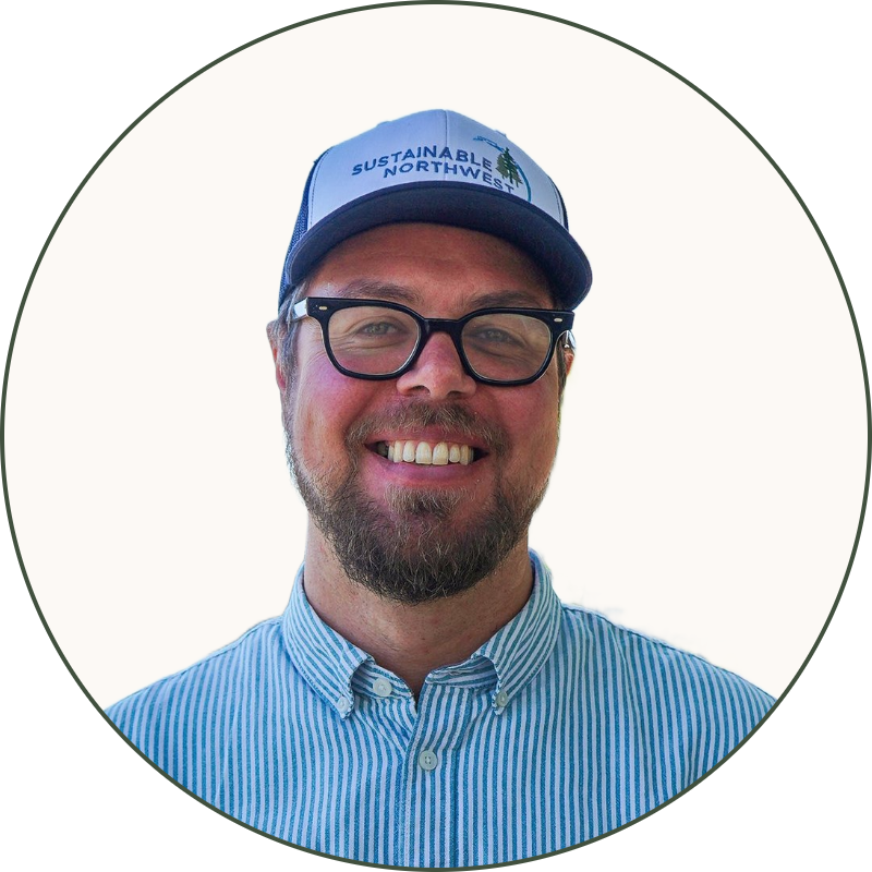 A smiling man wearing glasses, a blue and white striped button-up shirt, and a cap with the text 'Sustainable Northwest' standing outdoors near a river with a bridge in the background and green trees.