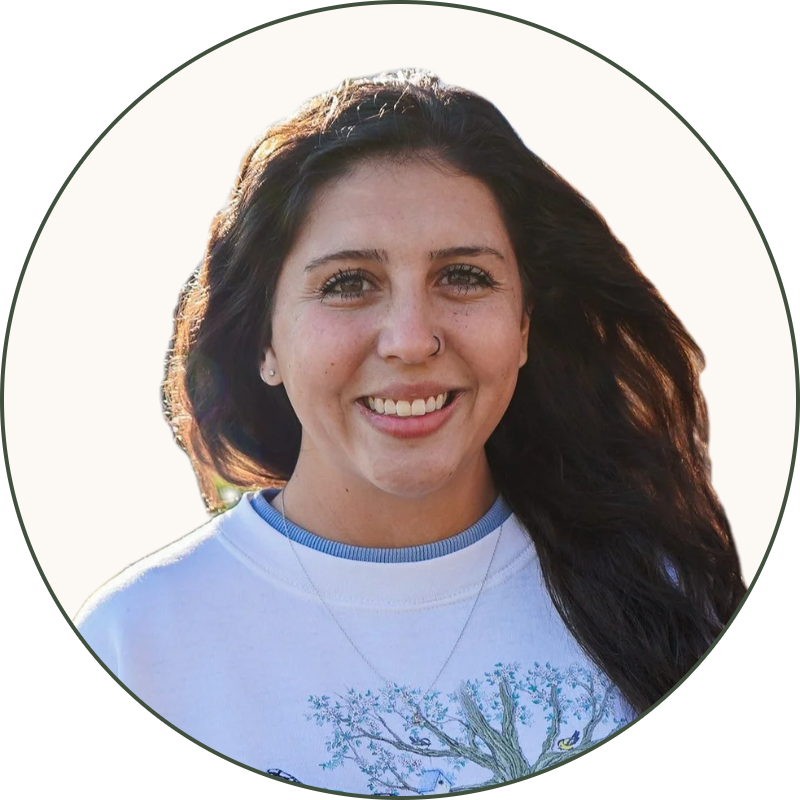 A young woman with long dark hair, smiling, wearing a white sweatshirt with a colorful butterfly and tree design, standing outdoors near a tree trunk with sunlight shining on her, in a park with trees and grass.