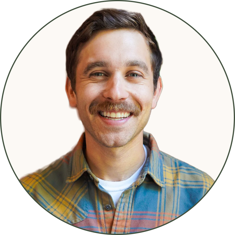 A smiling man with short dark hair, a mustache, and light skin, wearing a multicolored plaid shirt, standing in front of a red brick wall with a green plant to his right.