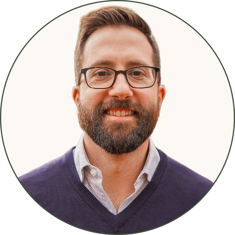 A smiling man with glasses and a beard standing outdoors on a fall day with trees and fallen leaves in the background.