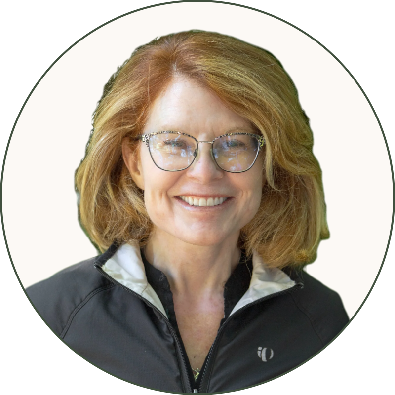 Close-up of a woman with red hair and glasses smiling, wearing a black jacket with a white collar, outdoors with blurred green foliage background.