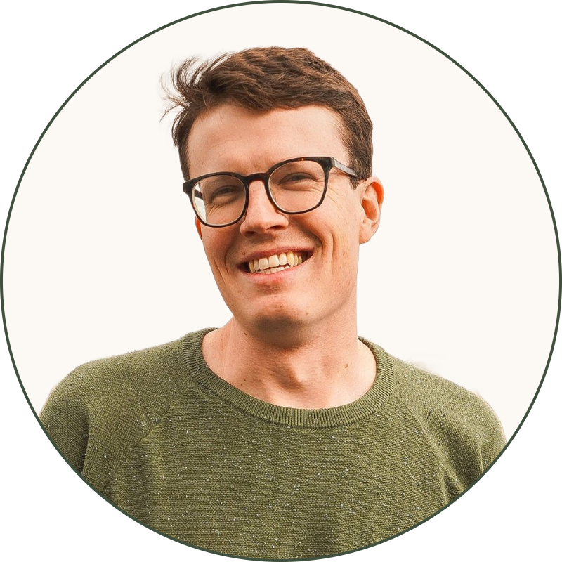 A young man with short brown hair, glasses, wearing a green sweatshirt and jeans, standing by a railing near a river, with an overcast sky in the background.