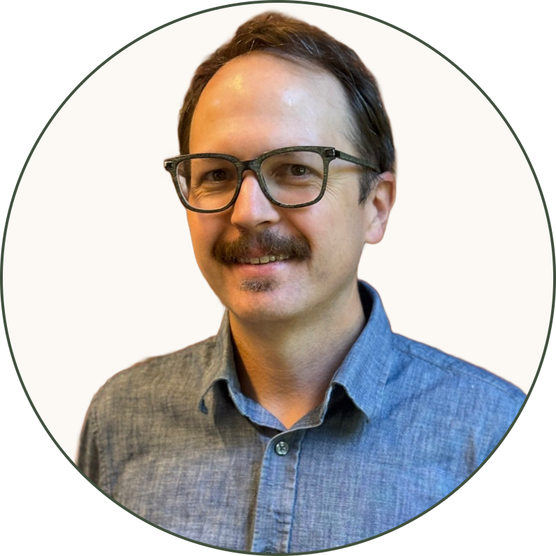 A man with glasses and a mustache wearing a collared shirt standing in front of a brick wall with a green plant to the right.