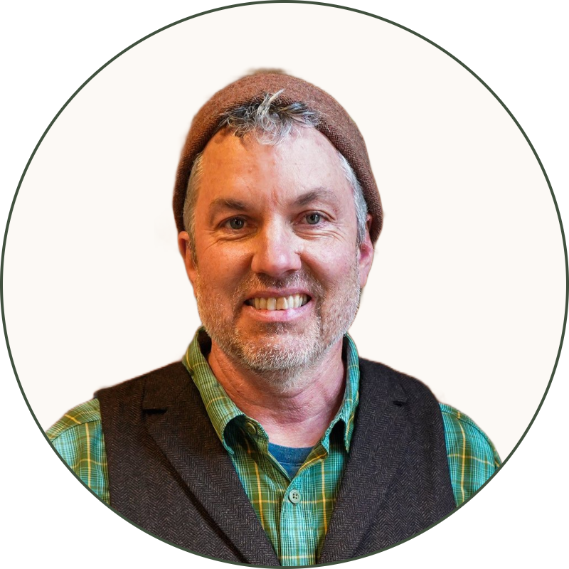 A man with gray hair and a beard wearing a brown hat, green plaid shirt, dark vest, and dark pants, standing in front of a brick wall and a tall indoor plant.