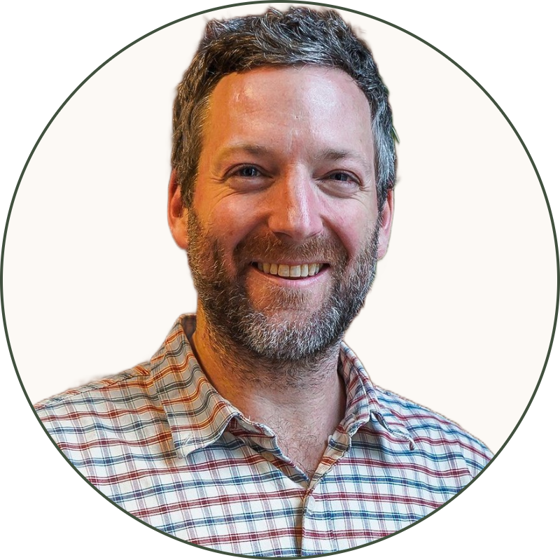 A smiling man with gray hair and a beard standing in front of a red brick wall and a green leafy plant. He is wearing a plaid shirt and jeans.