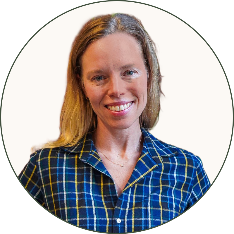A woman with blonde hair, wearing a blue plaid shirt and tan pants, standing in front of a brick wall with green plants.