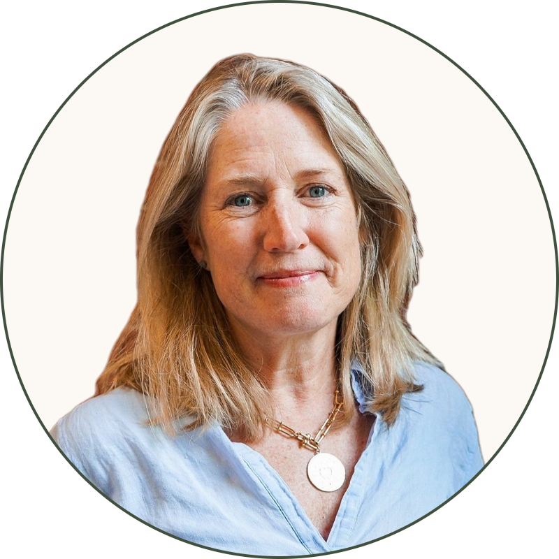 A middle-aged woman with blonde hair, light blue shirt, and blue jeans standing in front of a red brick wall with green plants on the right side.