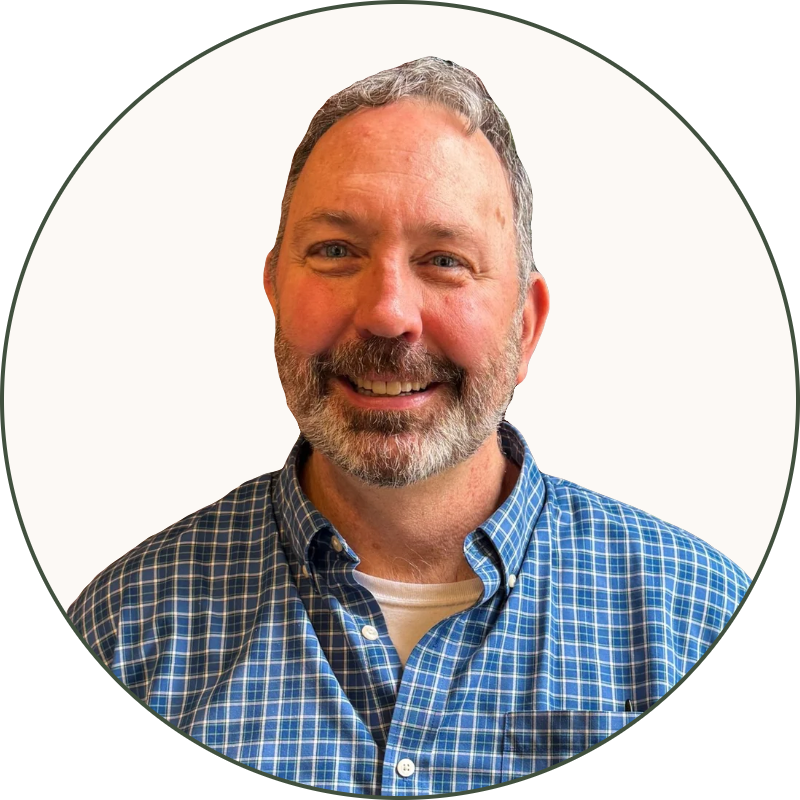 A smiling man with gray hair and beard wearing a blue checkered shirt standing in front of a brick wall and green leafy plant.