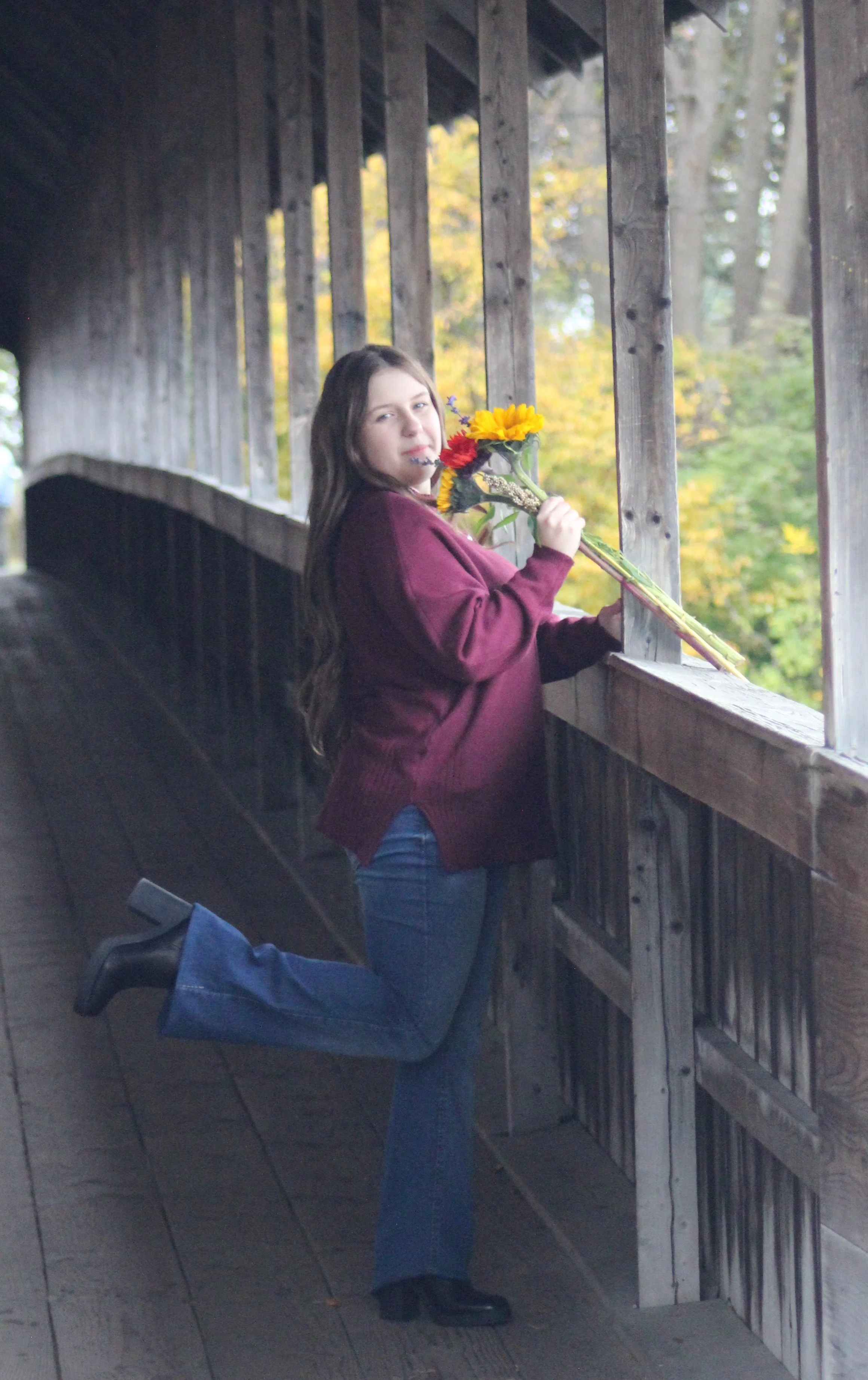 A woman with long brown hair wearing a maroon sweater and jeans kneeling on a wooden walkway, holding a bouquet of colorful flowers, with trees and autumn foliage in the background.