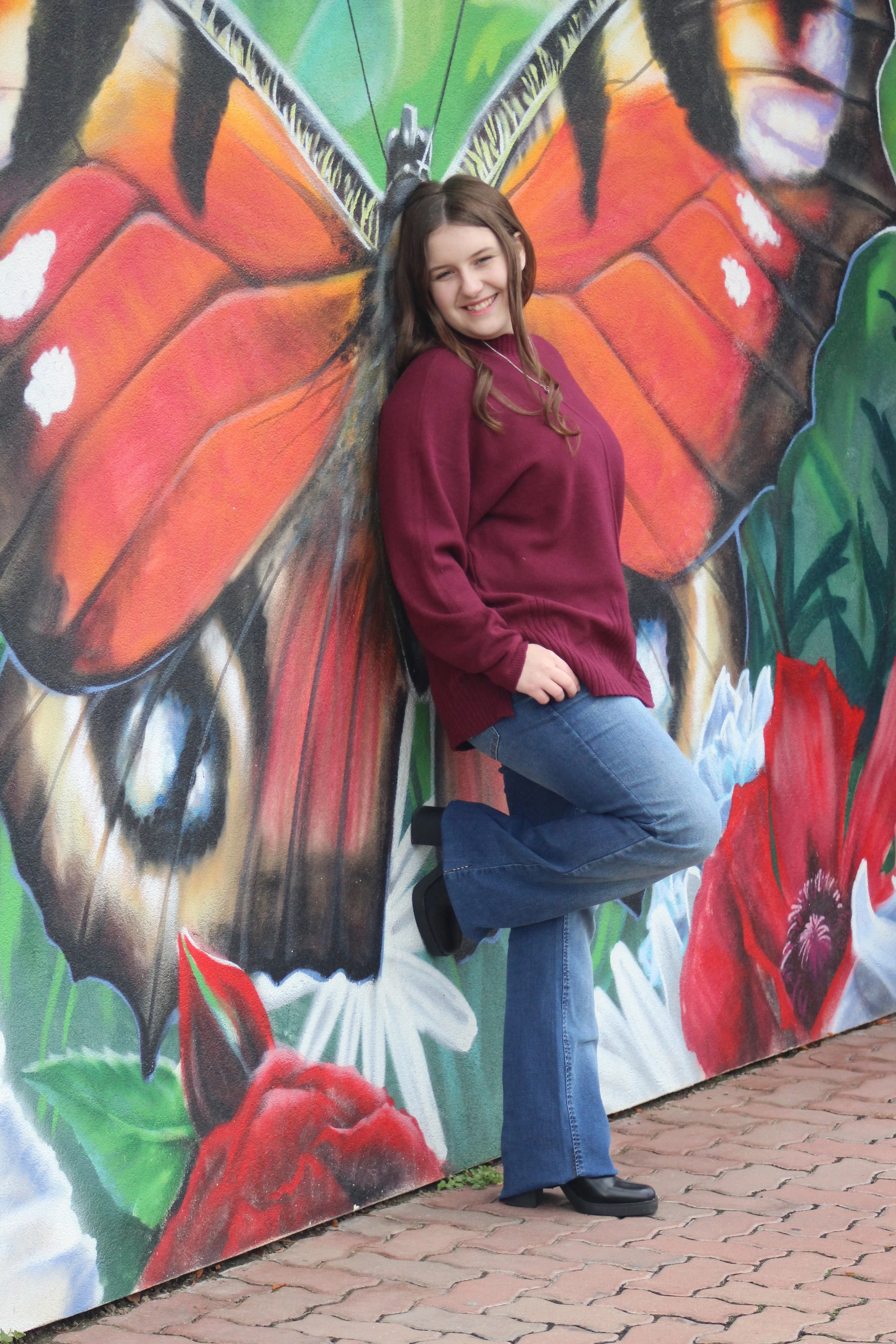 A young woman with long, wavy brown hair, wearing a maroon sweater, blue jeans, and black boots, smiling and posing with her back against a colorful butterfly mural on a wall. She is leaning against the mural with her eyes closed, her left arm restin