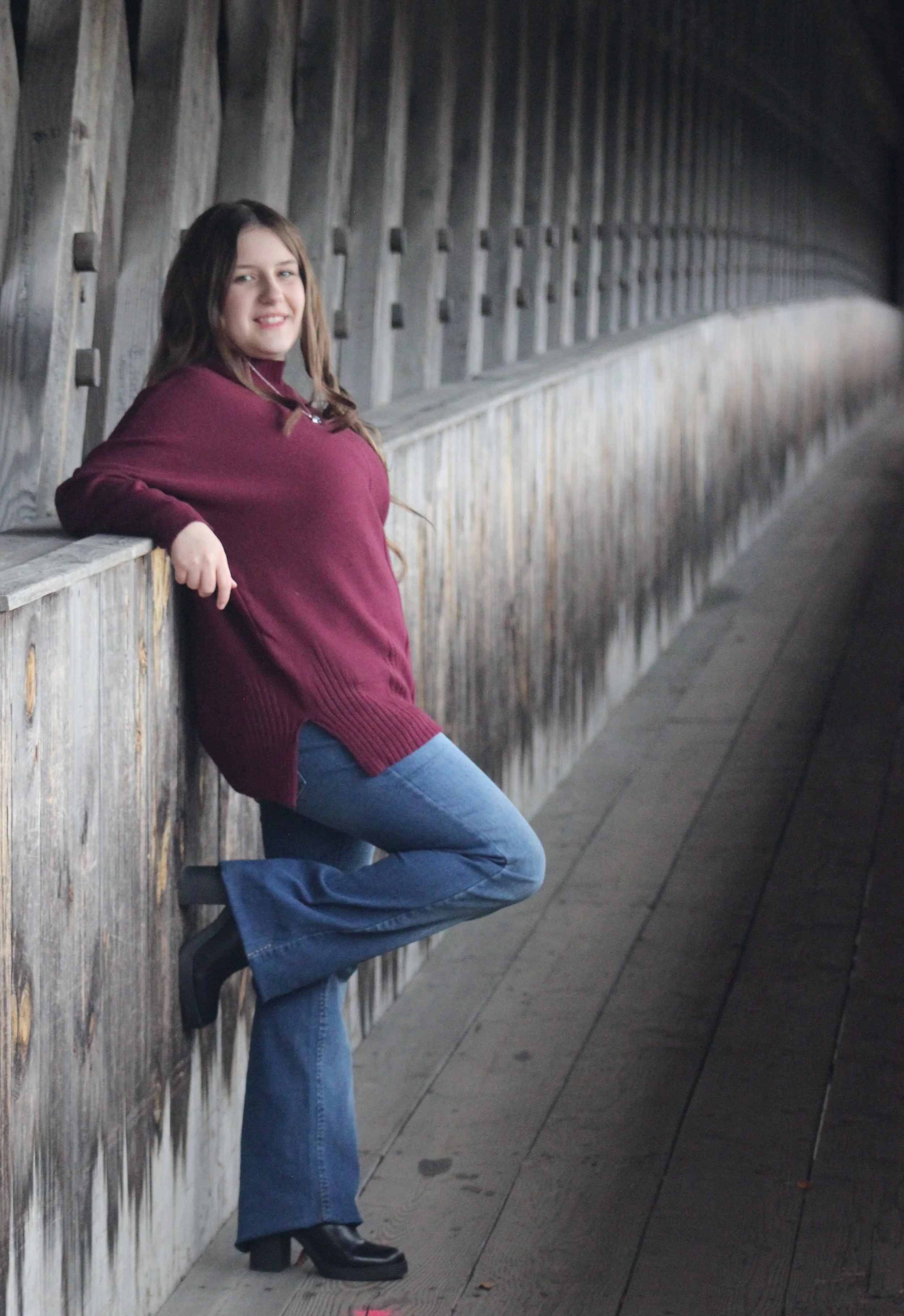 A woman standing beside a wooden fence in a tunnel-like structure, smiling at the camera, wearing a maroon sweater, blue jeans, and black boots.