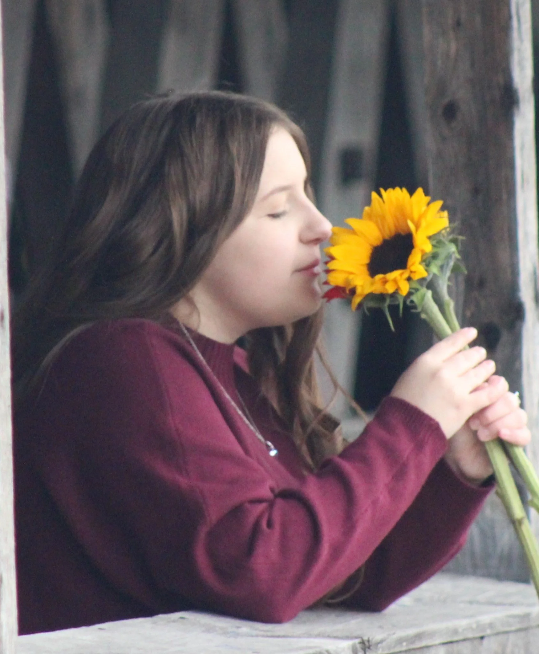 A woman with long brown hair holding a sunflower close to her face with her eyes closed, standing in front of a wooden background.
