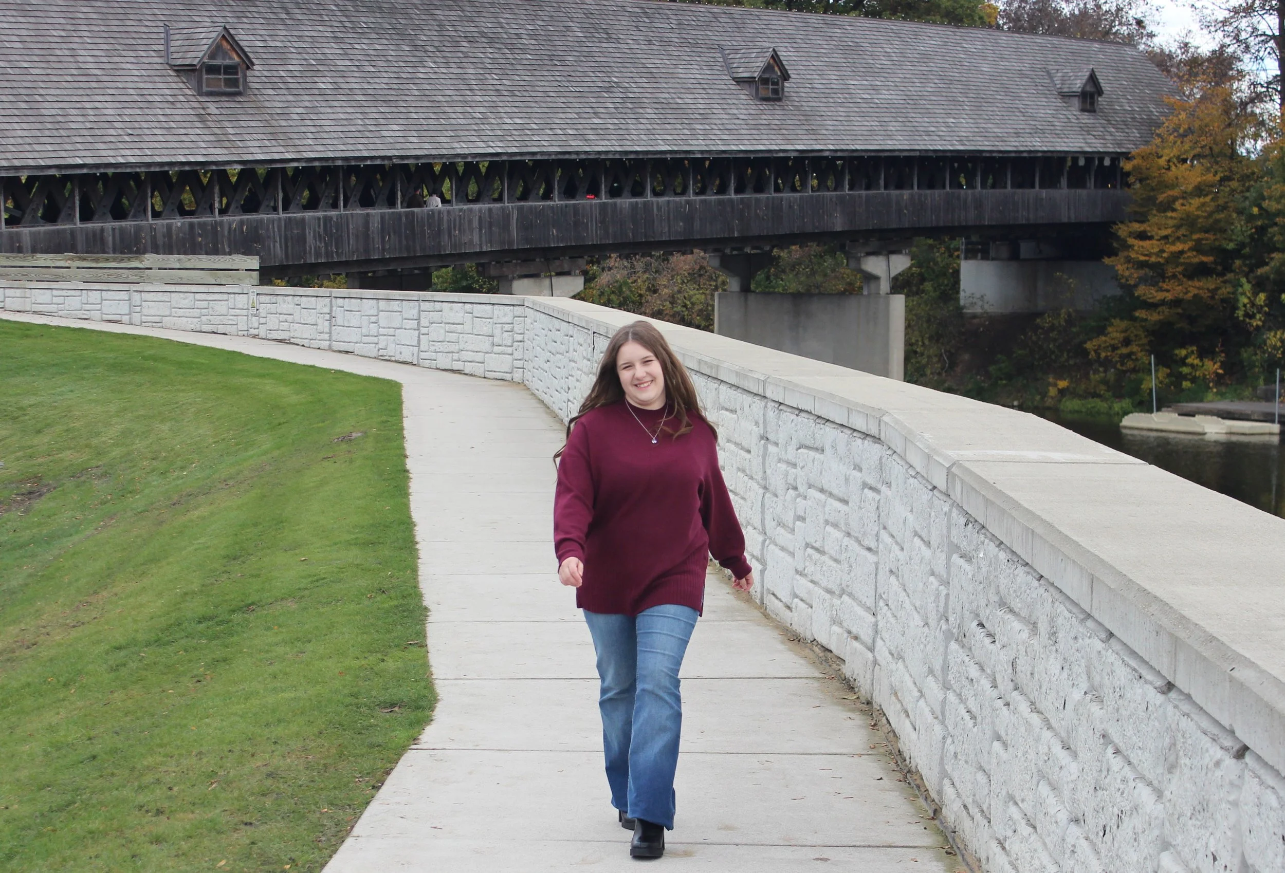 A young woman with long brown hair walking outdoors on a concrete path, smiling, wearing a burgundy sweater, blue jeans, and black boots, with a stone and wood bridge and green trees in the background.
