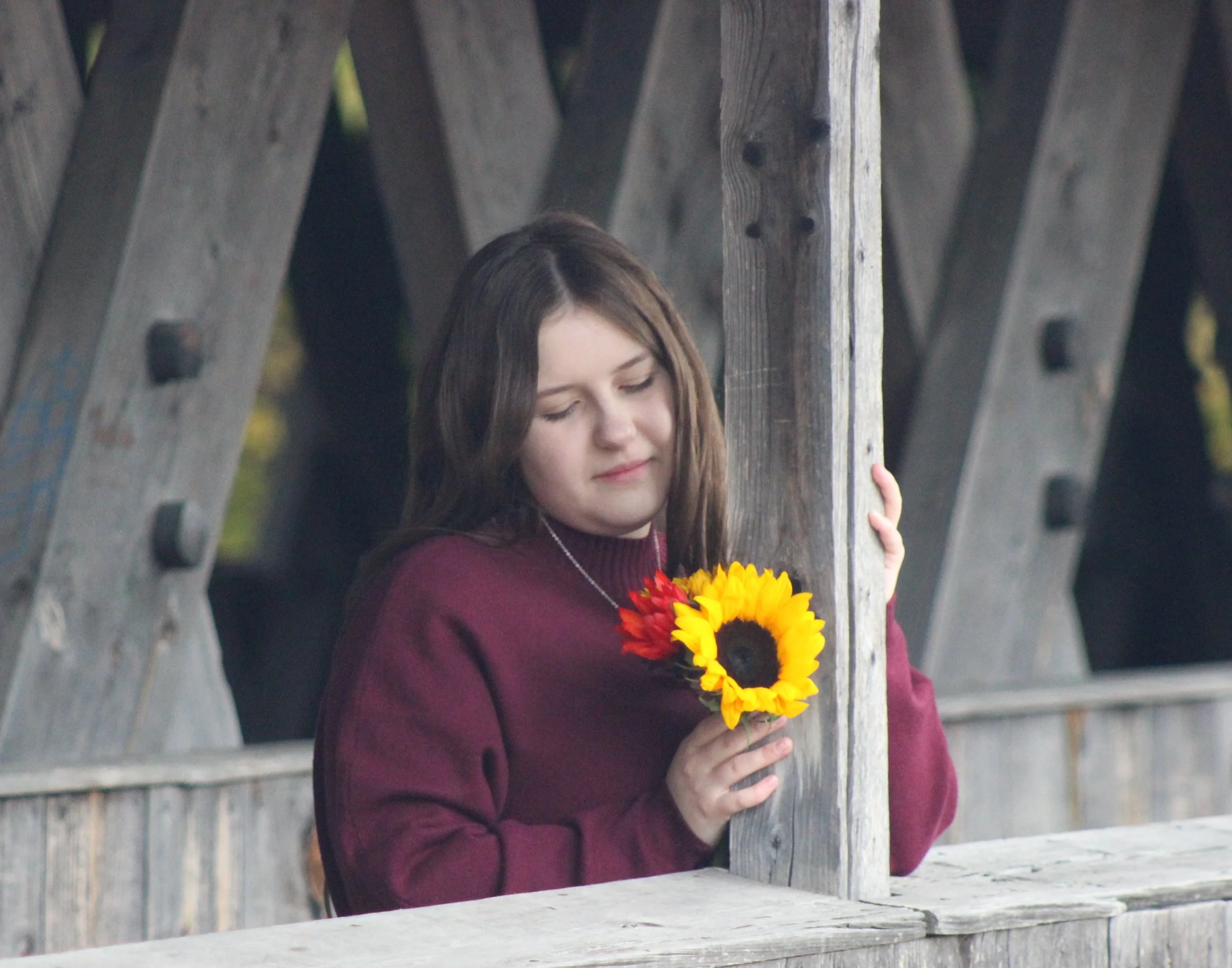 A young woman with long brown hair standing by a wooden fence holding a sunflower wreath and a bouquet of yellow and red flowers.