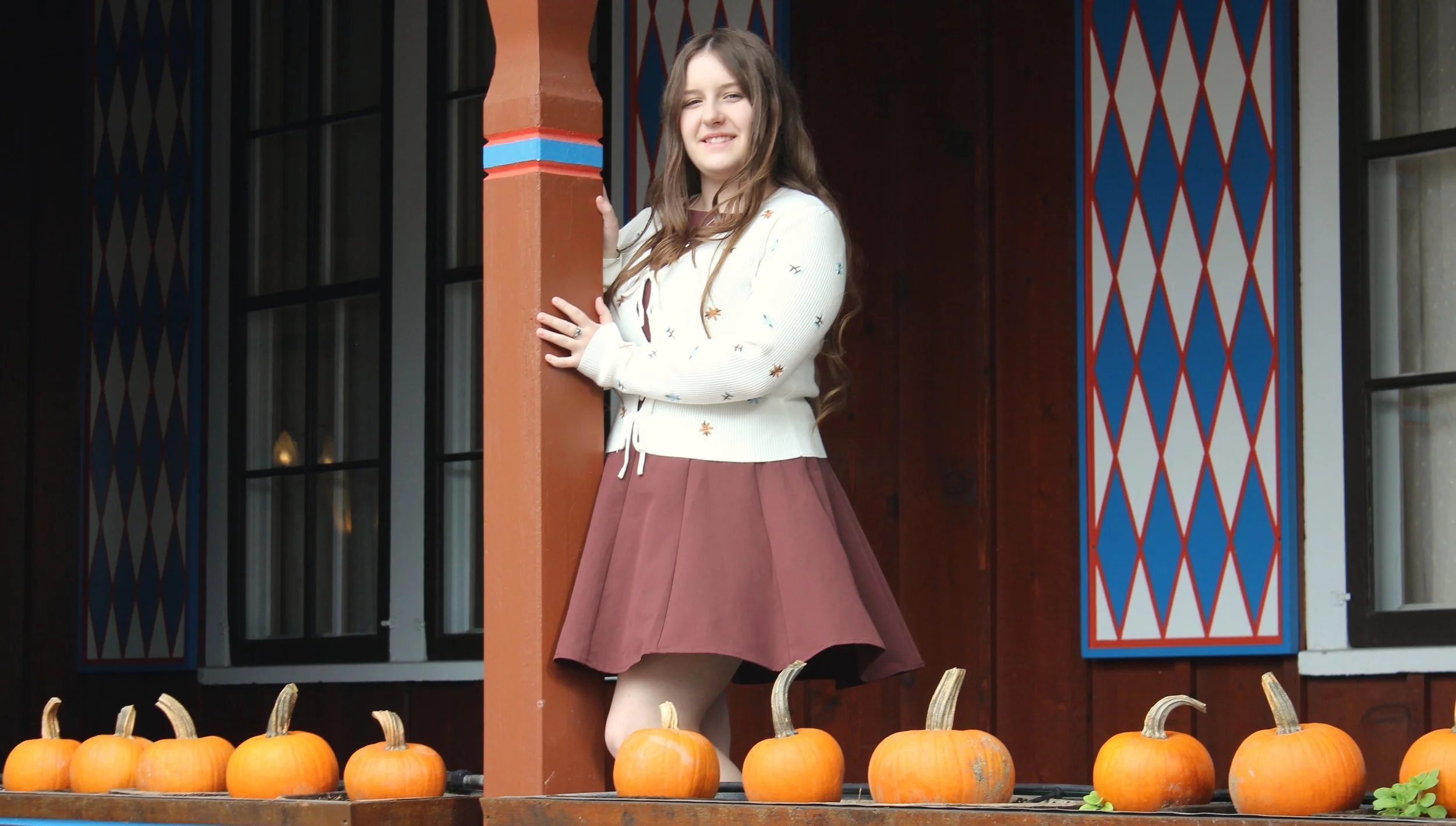 A young girl with long brown hair standing on a porch, partially behind a brown post, surrounded by orange pumpkins, with a background featuring a black window frame and colorful wall decorations in red, blue, and beige.