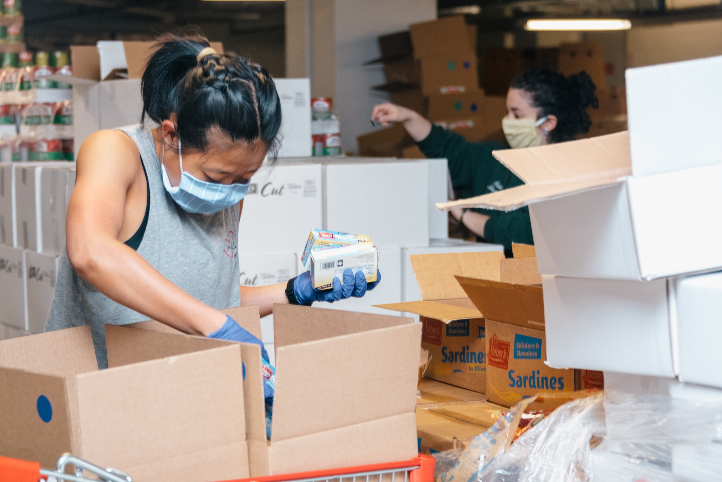 2 volunteers, each wearing face masks, putting cans of food into cardboard boxes at Met Council's Greenpoint fulfillment center.