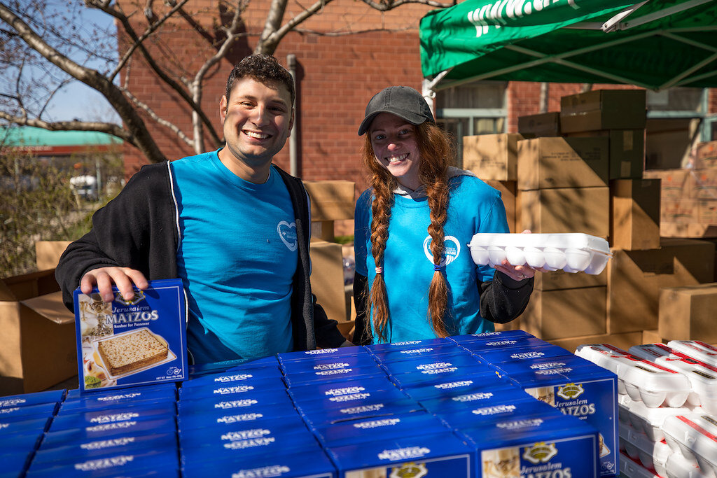 Two volunteers smiling at the camera in matching Met Council shirts at a Passover food drive. They are standing behind several boxes of Matzah on a table.