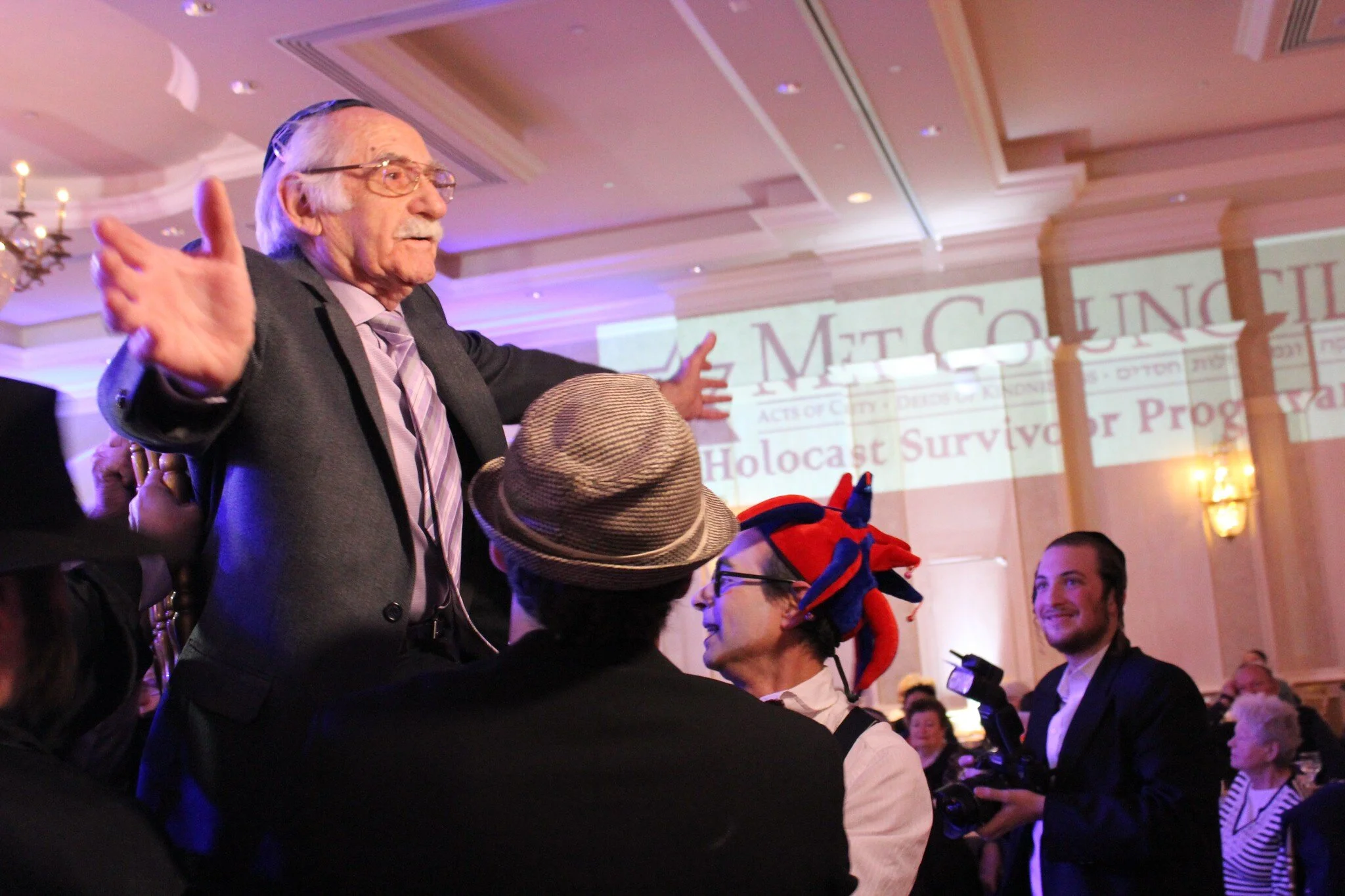 A holocaust survivor being held up on a chair at a party by a man with a tan fedora and a man with a red, felt jester’s hat.