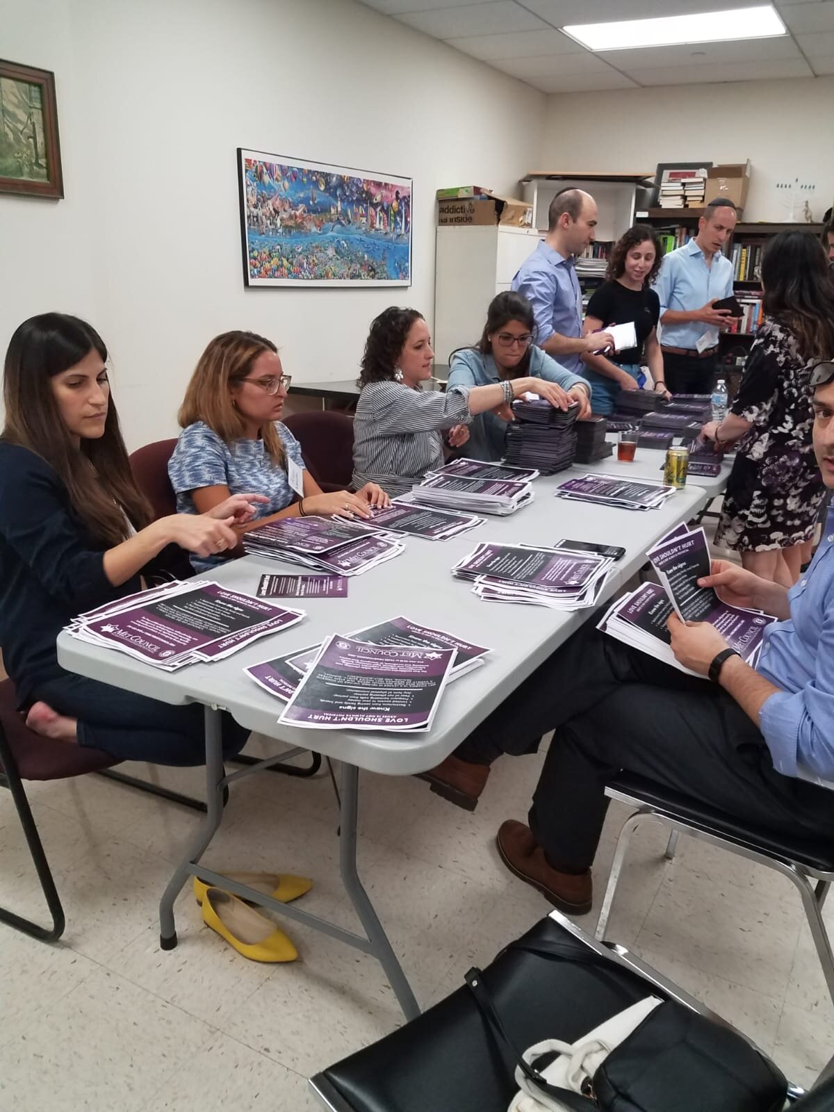 Volunteers sit at a long white table inside a rec room. They are all folding purple and black flyers for an event hosted by the Family Violence services program.