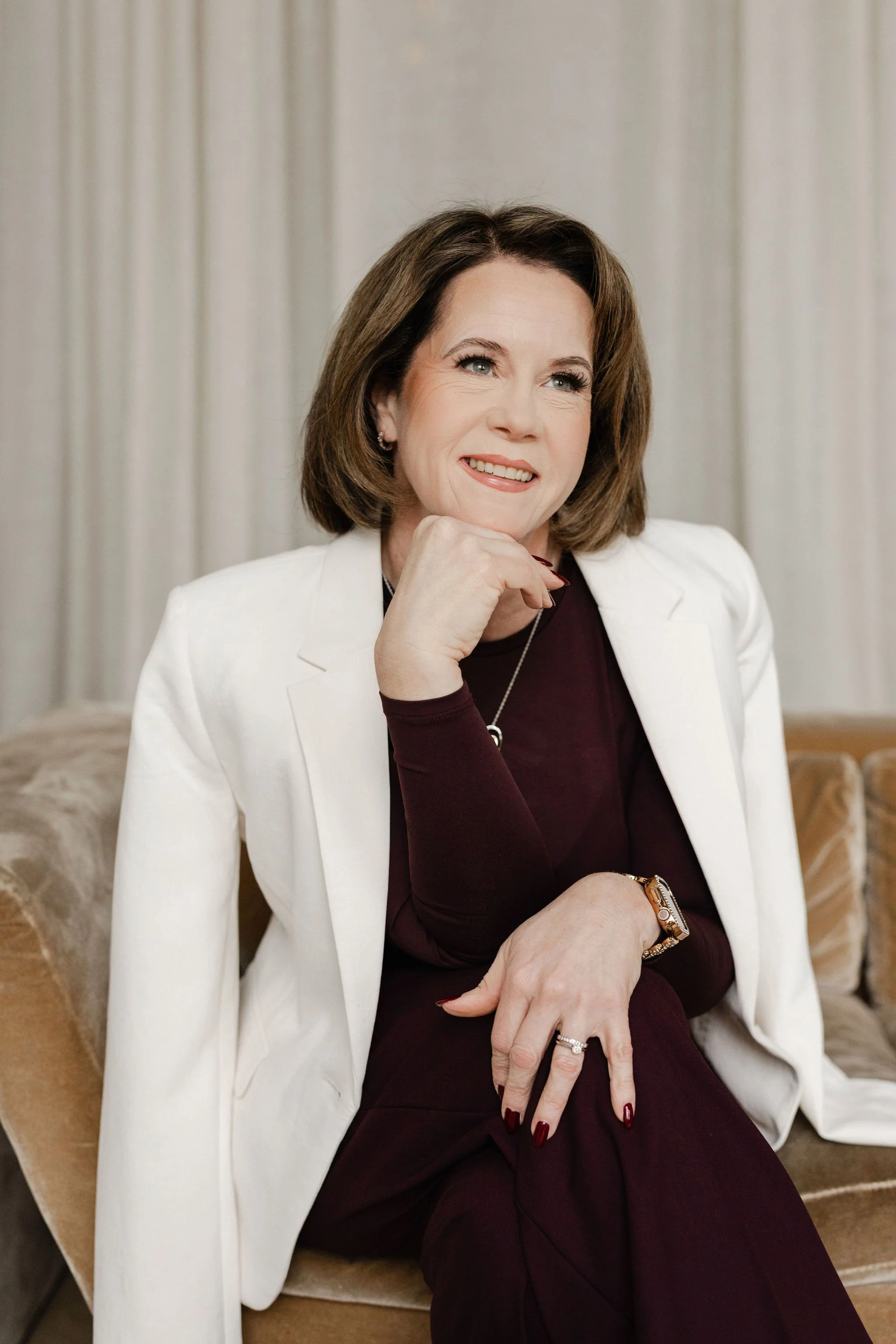 Vicky is in a pink blazer and floral top standing in front of black shelving with decorative items.