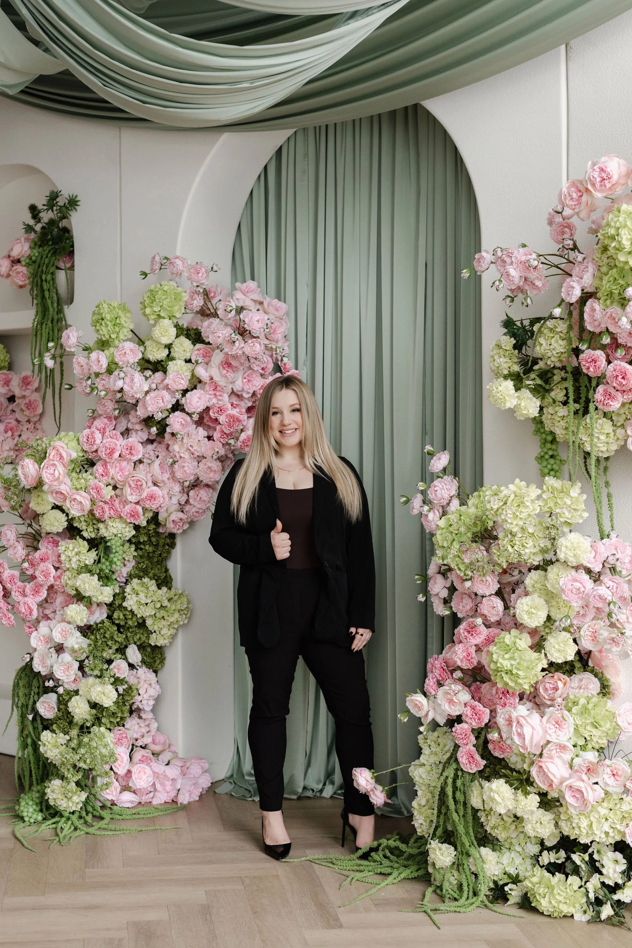 Cassandra, with long blonde hair wearing a black outfit stands in front of a pink brick wall with floral arrangements featuring pink and white flowers.