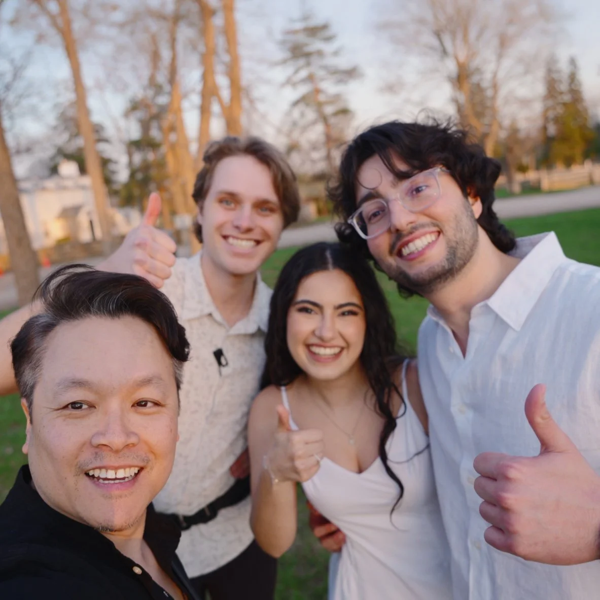 Group of five friends smiling and taking a selfie outdoors in a park during sunset, with trees and a grassy area in the background.
