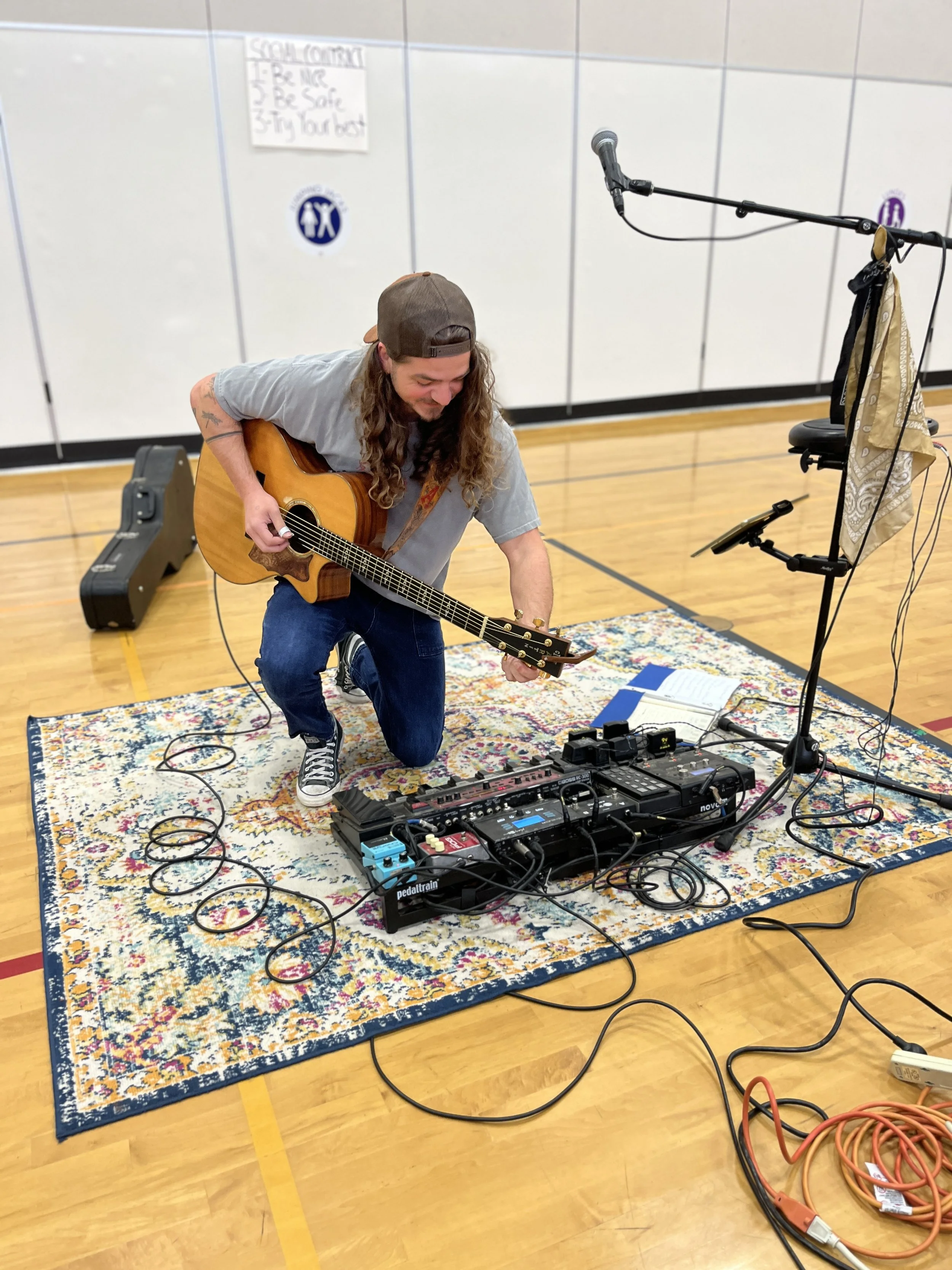 Musician with long hair, kneeling on a colorful rug, playing an acoustic guitar in a gymnasium with electronic music equipment and a microphone stand nearby.