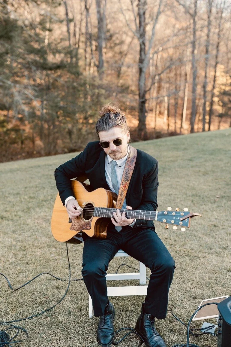 A man wearing sunglasses and a black suit playing an acoustic guitar outdoors on a grassy field with trees in the background.