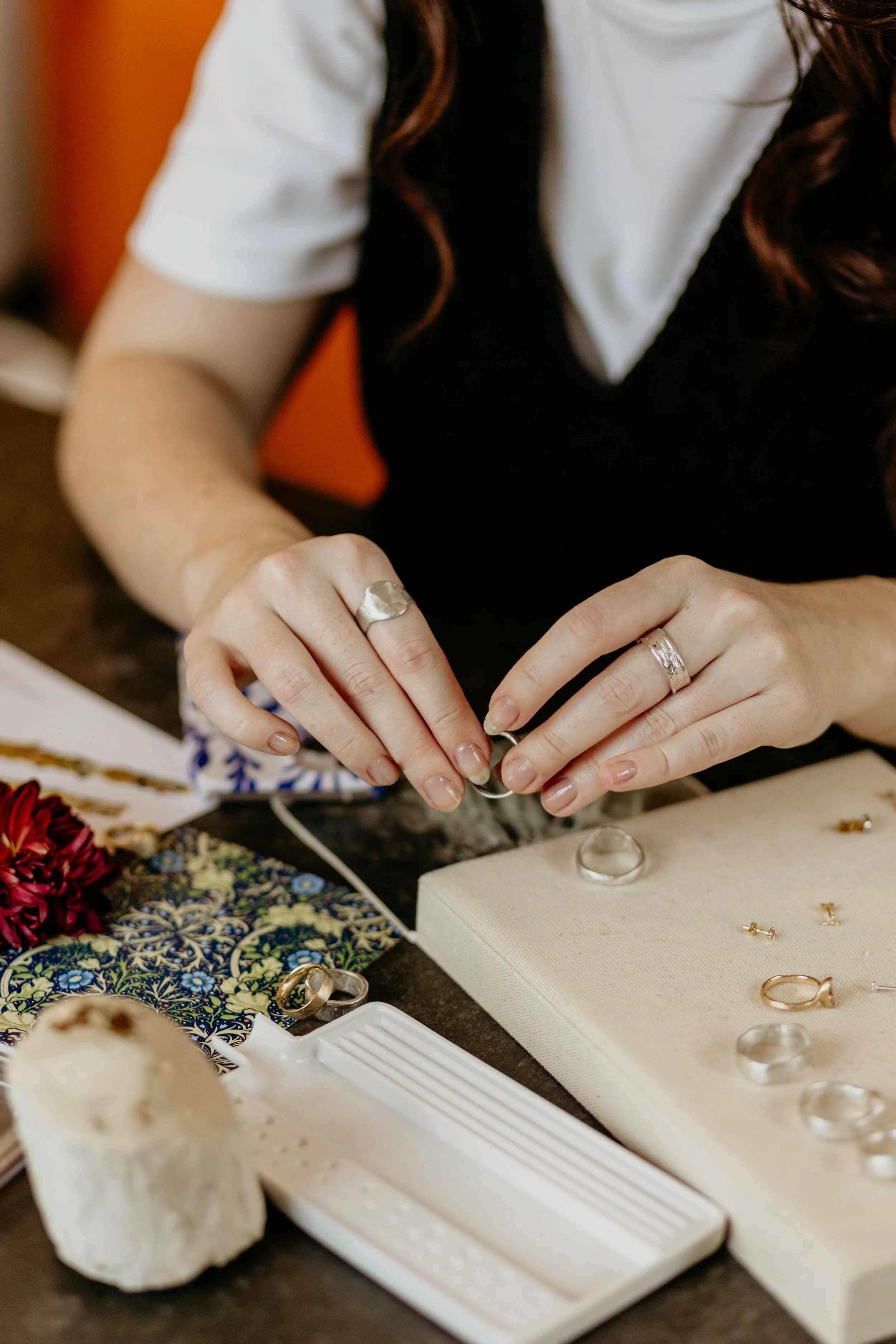 Close-up of a person working with jewelry, using hands to handle a ring, surrounded by jewelry pieces, fabric, and tools on a table.