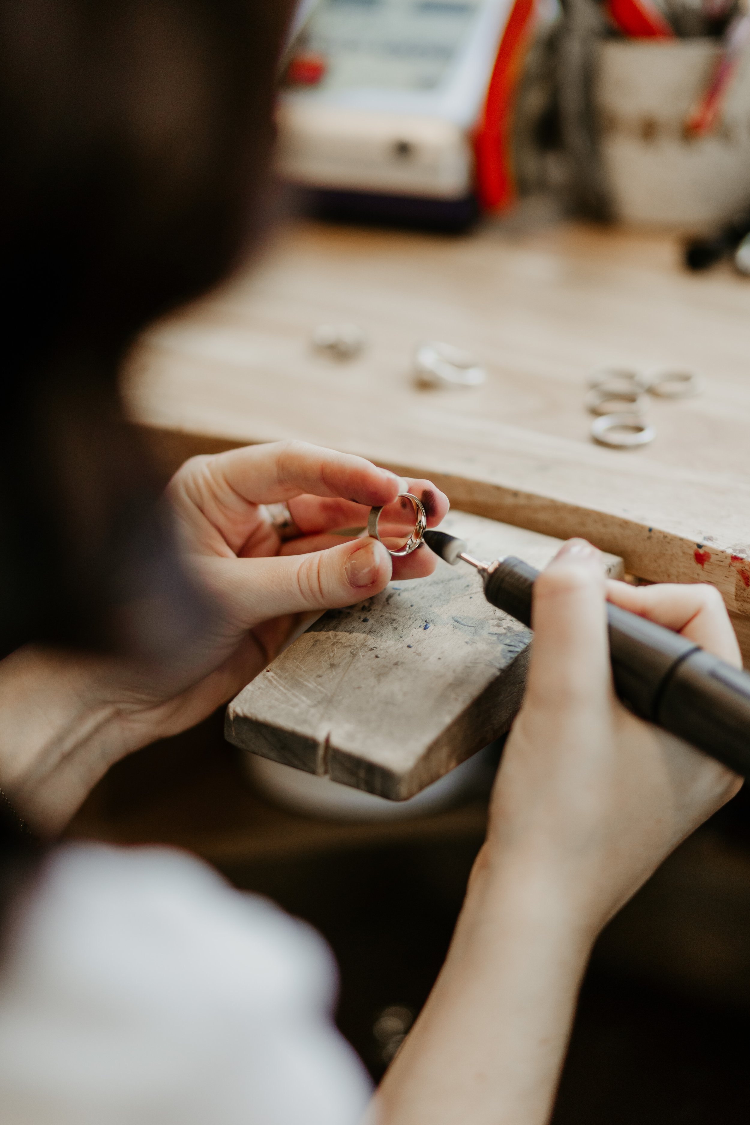 A person working at a craft or jewelry-making station, holding a ring and using a tool to modify or engrave it with small metal rings and jewelry tools on the wooden work surface.