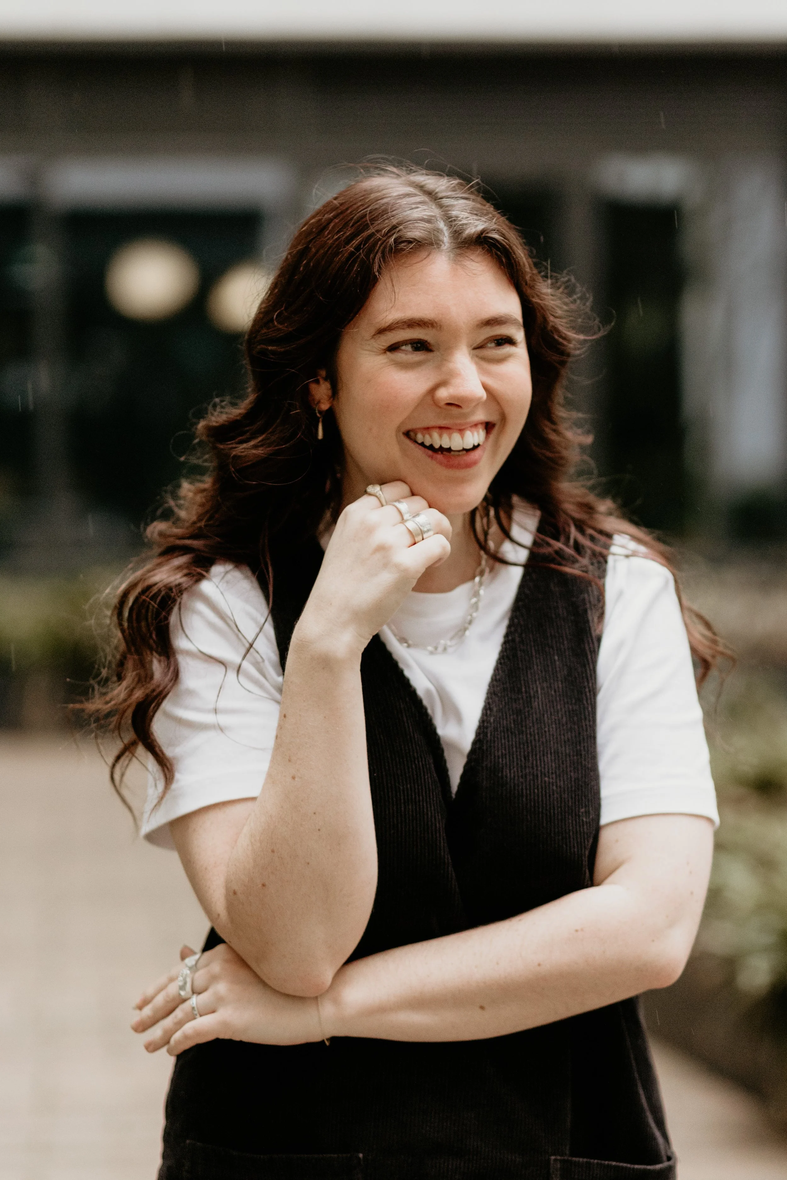 A young woman with long wavy brown hair smiling outdoors, wearing a white t-shirt layered with a black sleeveless top, with jewelry including rings and a necklace.