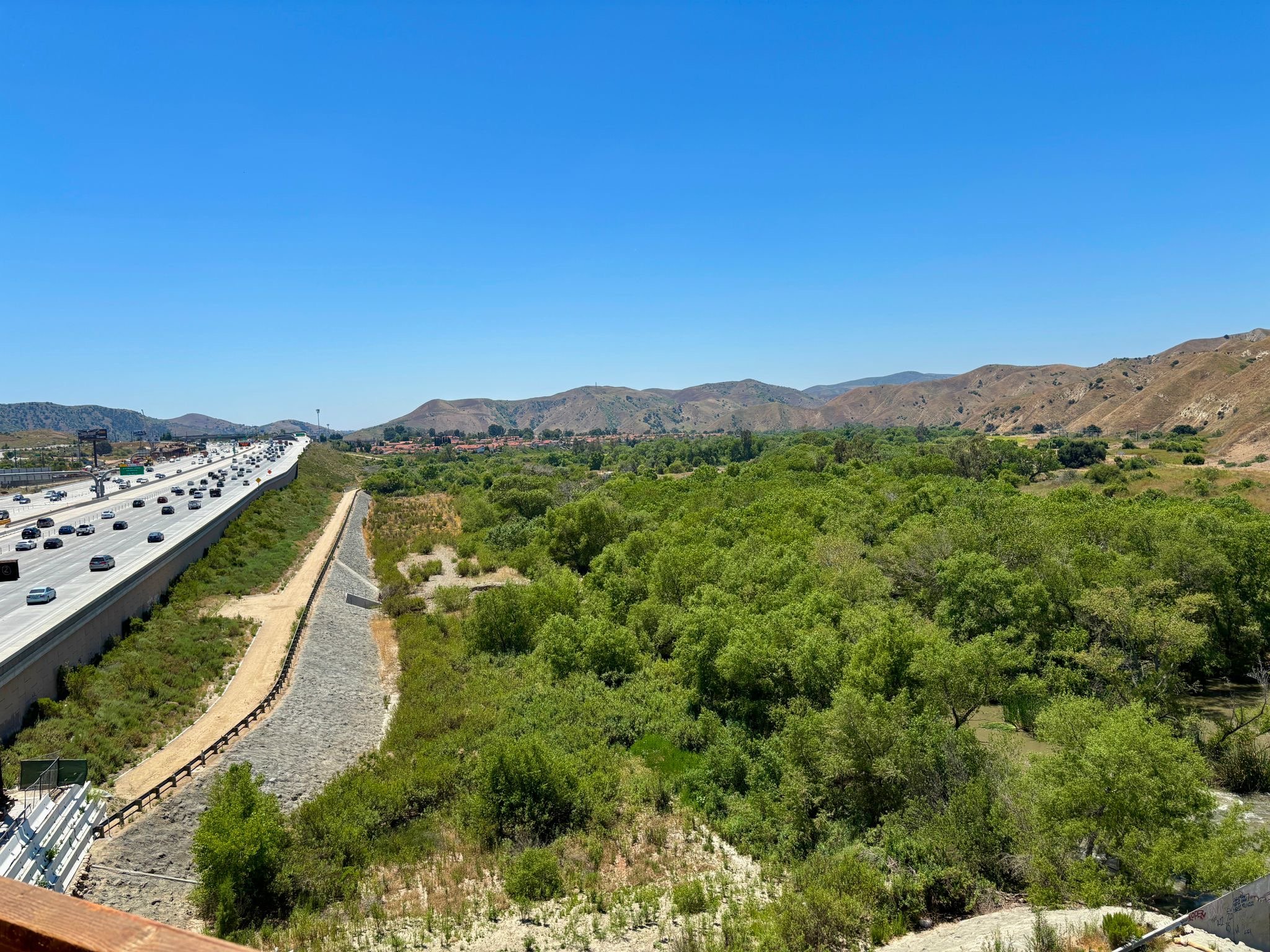 A highway with multiple lanes of traffic on the left side of the image, next to a green, tree-filled valley with mountains in the background under a clear blue sky.