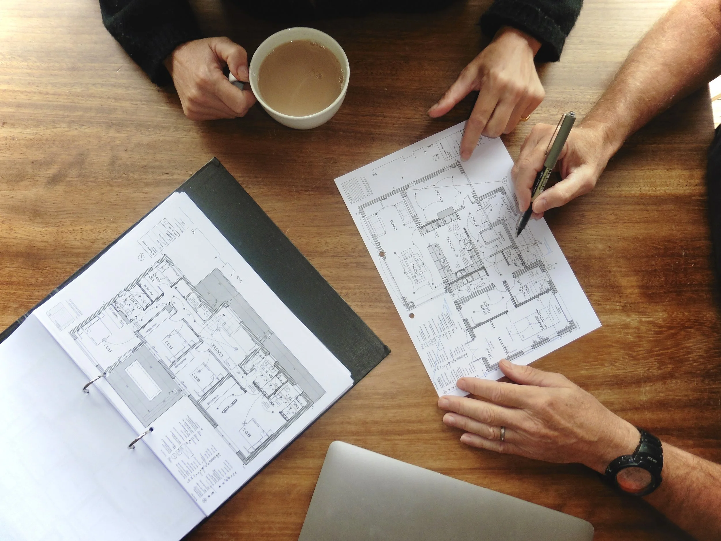 Two people are working on architectural blueprints at a wooden table, with one person pointing to a specific area on the blueprint and the other holding a coffee mug.