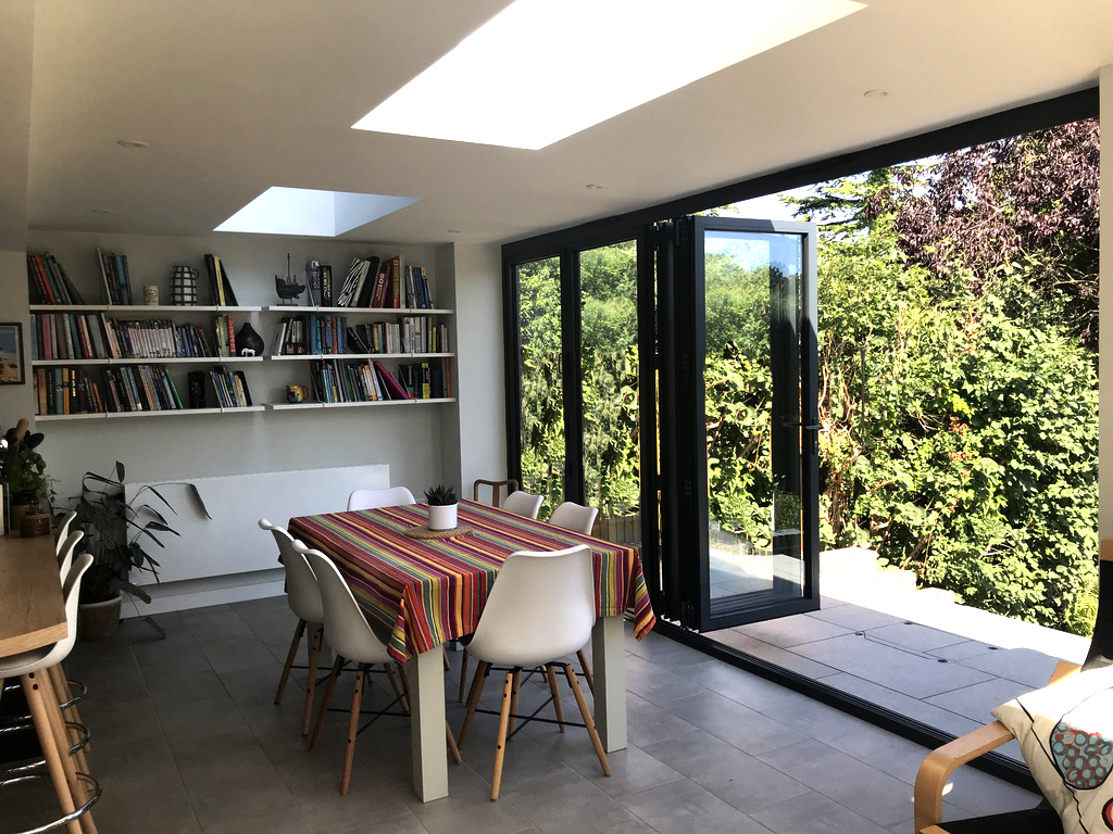 Bright dining area with a table covered in a colorful striped tablecloth, surrounded by white chairs. A large glass door opens to a lush green garden. Shelves filled with books and decorative items are on the wall.