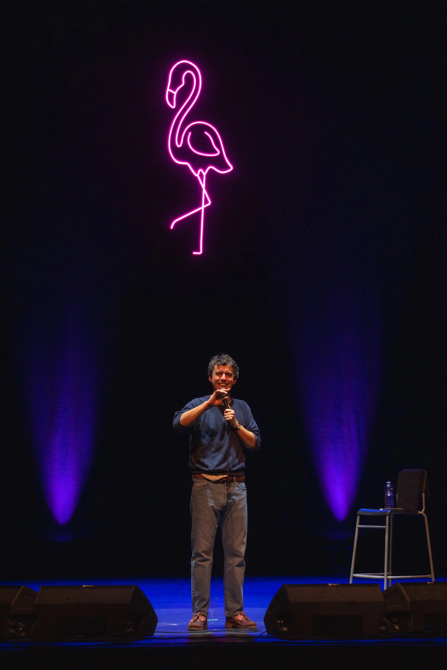 A man on stage holding a microphone, smiling, with a neon pink flamingo sign hanging above him and stage lighting.
