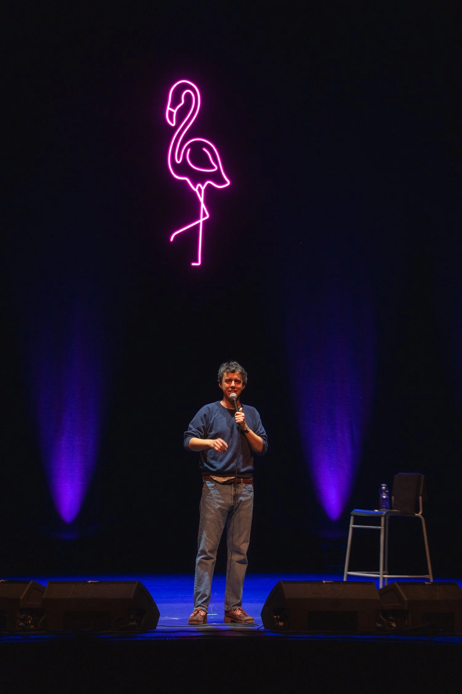 A man holding a microphone on stage with a neon pink flamingo sign above him, illuminated purple lights illuminate the background, and a chair with a water bottle is nearby.
