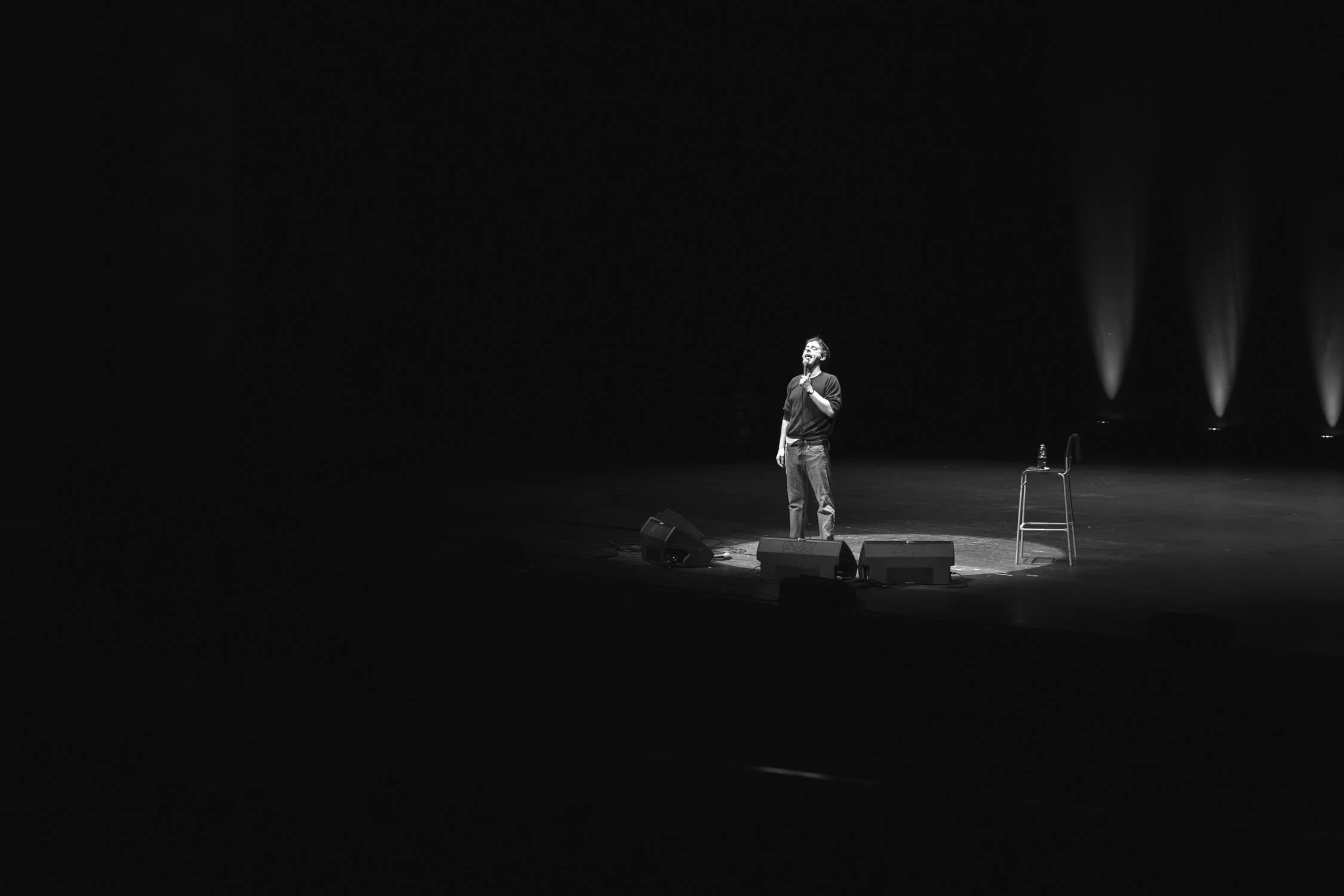 A person standing on a stage with a microphone, illuminated by a spotlight, with four spotlights in the background, a chair, and two water bottles nearby.